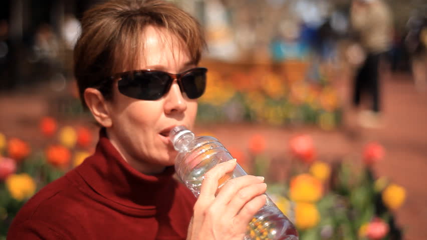 A woman drinking water outside a busy shopping mall with large flower beds full of multicolored tulips in the background.