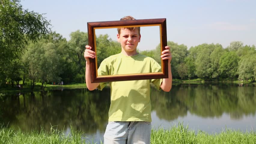 Boy in green t-shirt raises picture frame and shifts it sideward several times, near pond at summer park
