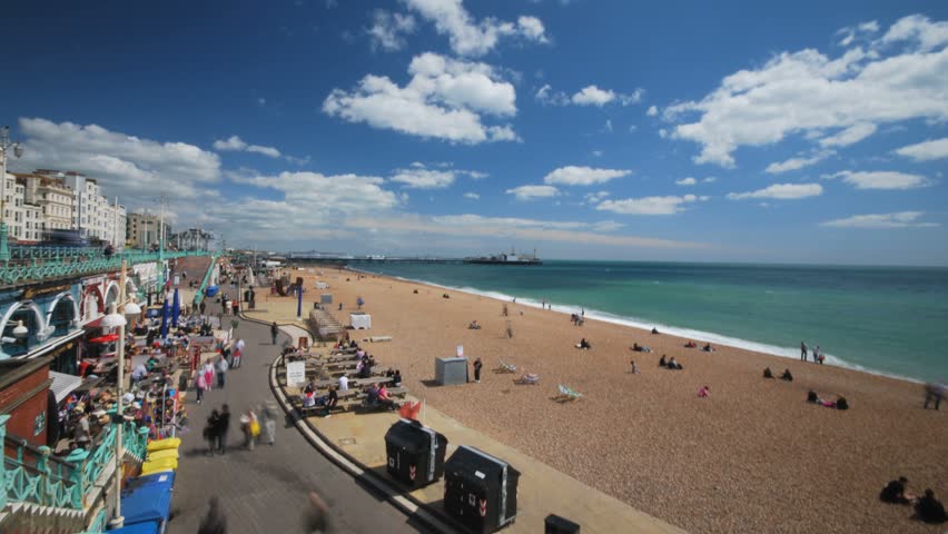 Time lapse of blue sky and clouds moving across a pebble beach and sea at Brighton pier as tourists enjoy the sun