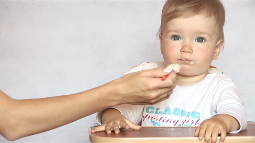 Baby sits at a children's little table. Mum feeds the child with porridge. Isolated on white.