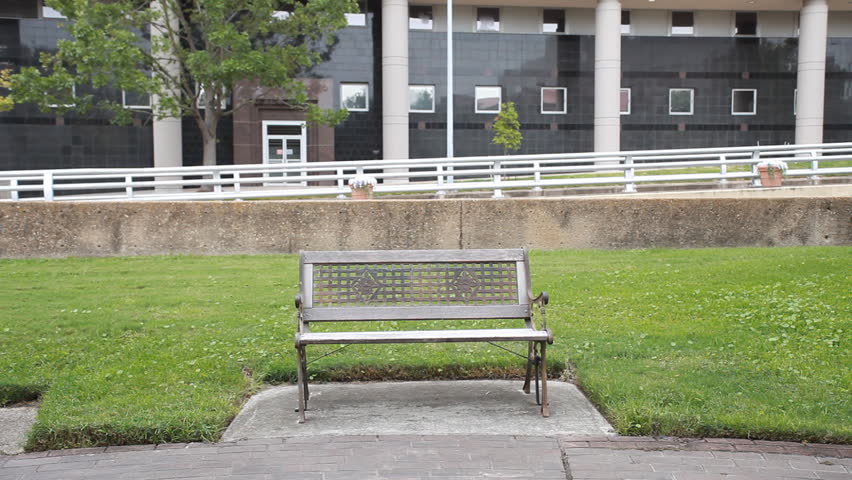A downcast man carrying a box walks over to a park bench, sits down, pulls a piece of paper out of a box and starts reading it.