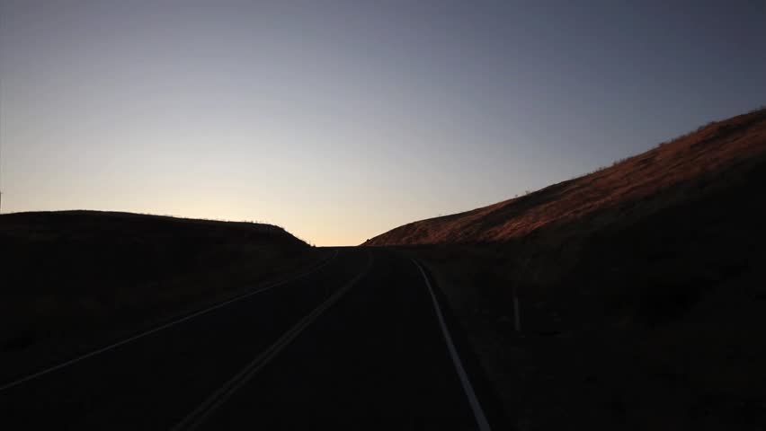 Camera cresting a hill from high vehicle point-of-view into rising sun and view of San Luis Reservoir and rolling hills in California with circular sunburst.