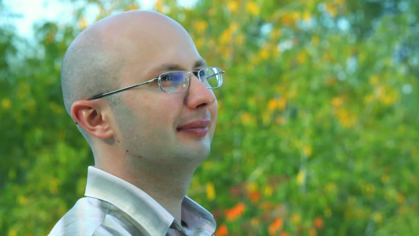 portrait of smiling bald-headed bespectacled man stands against trees in park 