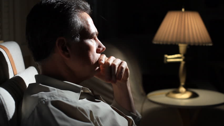 A man sitting in a dimly lit room, who appears preoccupied or troubled.
