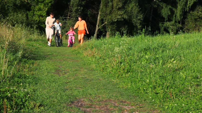 family of four, parents helps a kids riding bicycles to camera in park 