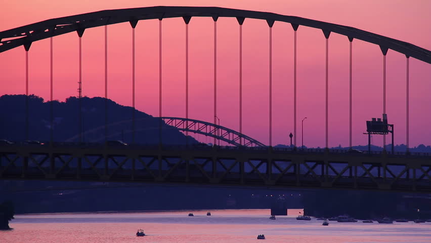 Traffic passes over the Fort Pitt Bridge during dusk in Pittsburgh, PA as pleasure boats travel underneath.