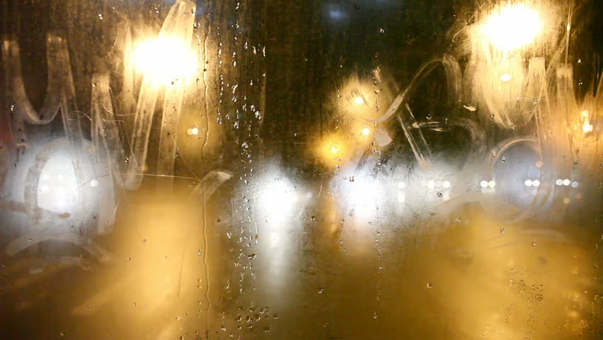 Steamy rear window view. Red traffic lights are left behind. Cold weather cityscape. Night time. Focus on window. Glowing blurred lights. 