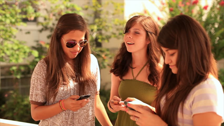 Teenage girls standing outside on a bright sunny day using their phones to send text messages to their friends.
