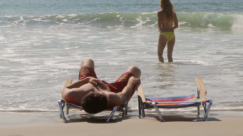 Woman walking out of ocean to boyfriend laying in lounge chair 