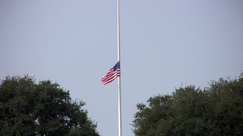Video zoom out of the Fort Sam Houston Veterans Cemetery. Headstones and flag at half staff. Green grass and oak trees in distance. Memorial to those who served the Armed Forces. San Antonio, Texas. 