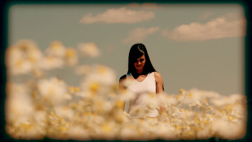 Young woman walking in the meadow, touching camomiles and smiling at camera stylized at reel movie