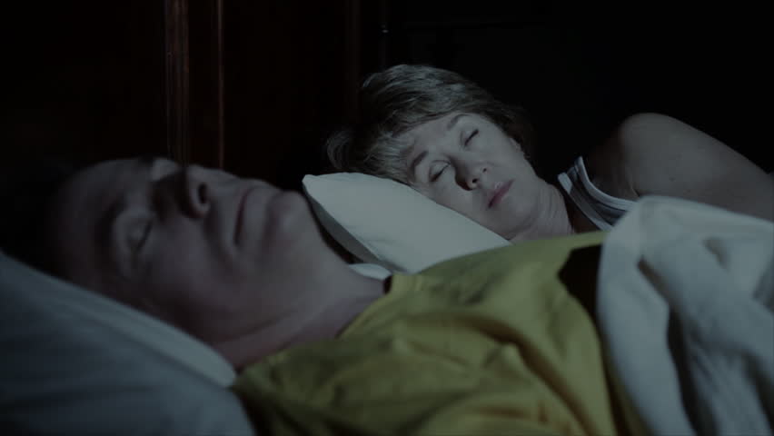 A sleepless woman lies wide awake in bed while her husband is sleeping soundly next to her.