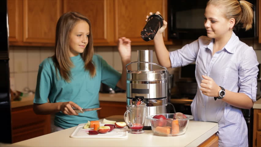 Two adolescent girls standing in a kitchen have fun adding fruits and vegetables to a juicer to make a nutritious vitamin packed drink.