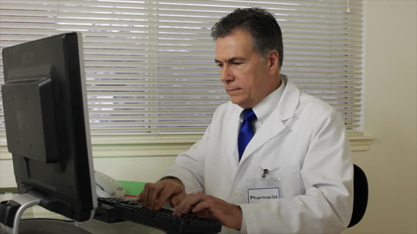 A pharmacist working at his computer answers the phone and engages in conversation.