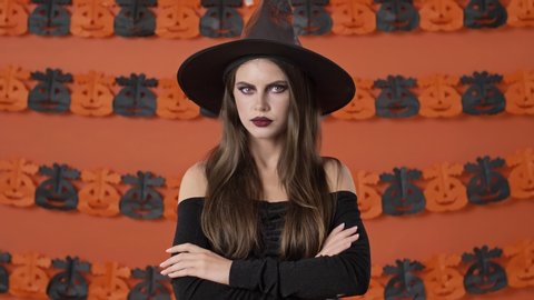 Serious Pretty Young Witch Woman In Black Halloween Costume Saying No And Shaking Her Head Negatively Over Orange Pumpkin Wall