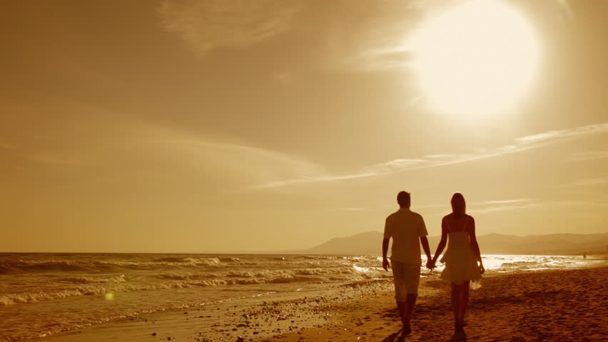 Senior Adult Couple Holding Hands Walk Up The Beach As The Sun Is About To Set On The Horizon 